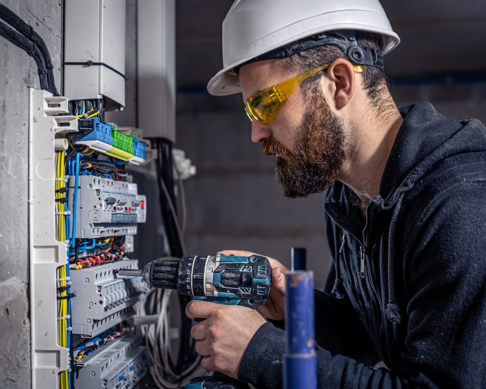 A male electrician works in a switchboard with an electrical connecting cable, connects the equipment with tools.